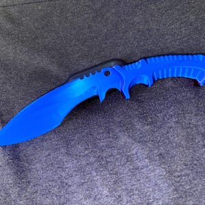 May include: A vibrant blue knife with a curved blade and a textured handle. The knife features a black section near the handle and is set against a dark grey background. The blade is sharp and the handle has finger grooves.