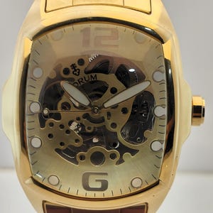May include: A gold-toned wristwatch with a tonneau-shaped case and a matching link bracelet. The dial features a skeletonised design revealing internal mechanical components, white-tipped hands, the text CORUM, and large numerals 12 and 6.