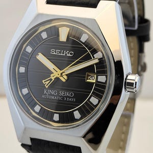 Könnte beinhalten: Eine King Seiko Automatikuhr mit schwarzem Zifferblatt mit horizontalen Linien, goldfarbenen Zeigern und weißen Stundenmarkierungen. Die Uhr hat ein silberfarbenes Edelstahlgehäuse und ein schwarzes, strukturiertes Lederarmband mit Datumsanzeige auf 3 Uhr.