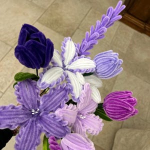 Puede incluir: Un ramo de flores hechas a mano en tonos morados, lavanda y blanco. Las flores incluyen tulipanes, flores en forma de estrella y una ramita de lavanda. Los tallos son verdes y las flores están hechas de un material suave y texturizado.