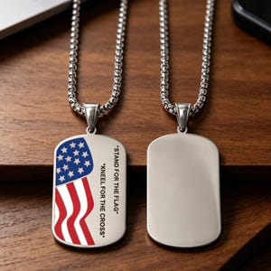 Könnte beinhalten: Zwei silberfarbene Erkennungsmarken-Anhänger an passenden Boxketten. Ein Anhänger zeigt eine aufgedruckte amerikanische Flagge sowie den Text "STAND FOR THE FLAG" und "KNEEL FOR THE CROSS". Der zweite Anhänger ist eine glatte, polierte silberfarbene Fläche.