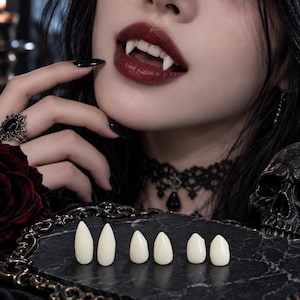 Könnte beinhalten: Ein Set aus sechs cremeweißen, spitzen Vampirzähnen in verschiedenen Größen, die in einer Reihe auf einer schwarzen Marmoroberfläche angeordnet sind. Die Zähne sind glatt, gebogen und so gestaltet, dass sie scharfe Eckzähne imitieren.