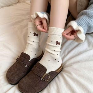 Könnte beinhalten: Cremefarbene Socken mit einem Muster aus kleinen schwarzen Punkten und braunen Dackel-Illustrationen. Dazu braune Wildleder-Clogs mit goldfarbenen Schnallen. Die Clogs haben eine Holzsohle.