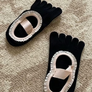 May include: A pair of black toe socks with a sandal-like design. Each sock features a pink strap and a white ruffled trim around the open area. The socks are designed to separate the toes.