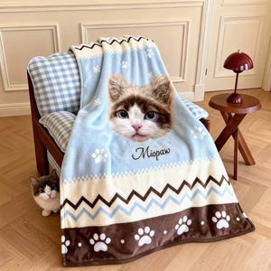 Könnte beinhalten: Hellblaue Decke mit einem Katzenmotiv. Sie zeigt ein Katzen-Gesicht, Pfotenabdrücke, Schneeflocken und ein Zickzackmuster. Das Wort "Miopaw" ist auf der Decke geschrieben.