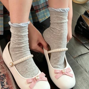 May include: White Mary Jane shoes with a pearl strap and a pink bow. The shoes are paired with gray socks with a bow pattern and a light blue trim.