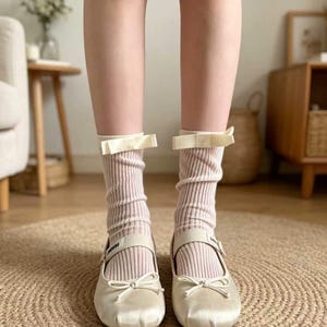 Könnte beinhalten: Hellrosa gerippte Socken mit cremefarbenen Schleifen, kombiniert mit cremefarbenen Mary Jane-Schuhen aus Satin mit Schleifendetails und Schnallenriemen. Die Socken und Schuhe befinden sich auf einem gewebten Teppich.