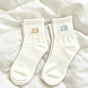 Könnte beinhalten: Zwei Paar weiße Knöchelsocken mit gerippten Bündchen. Ein Paar hat goldfarbene gestickte Initialen "J&M", das andere hat blau gestickte Initialen "J&M". Die Socken werden auf einer weißen, strukturierten Oberfläche präsentiert.