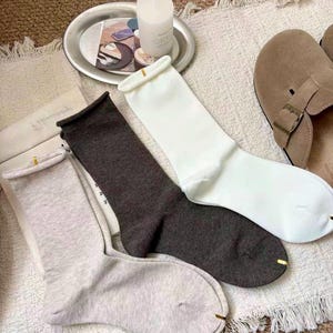 Könnte beinhalten: Drei Paar Socken in Hellgrau, Dunkelgrau und Weiß, jeweils mit einem goldenen Akzent an der Spitze. Die Socken sind gefaltet und auf einem strukturierten, cremefarbenen Teppich angeordnet. Ein silbernes Tablett mit einer Kerze und dekorativen Gegenständen befindet sich im Hintergrund.