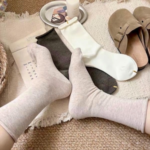 May include: A collection of socks in various neutral colors, including beige, white, and gray, are arranged on a textured surface. A pair of brown suede slippers and a candle on a silver tray are also visible. The socks appear to be made of a soft, comfortable material.
