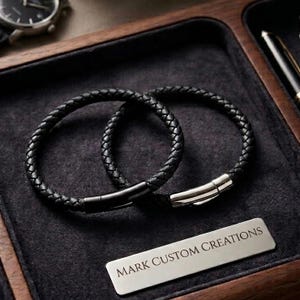 Könnte beinhalten: Zwei schwarze geflochtene Lederarmbänder mit silbernen Verschlüssen werden auf einer dunkelgrauen Oberfläche präsentiert. Die Armbänder sind kreisförmig und haben eine gewebte Textur. Ein silbernes Namensschild trägt die Aufschrift "MARK CUSTOM CREATIONS".