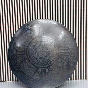 May include: A large, circular, dark metal shield with intricate engraved Celtic knot and floral patterns. The shield has a central design with radiating segments and a detailed border. The background features a striped pattern.