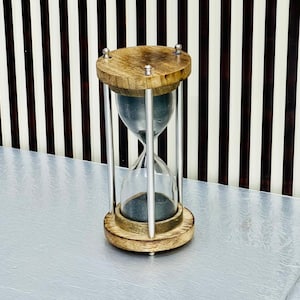 Peut inclure: Un sablier avec un dessus et une base en bois, reliés par des tiges métalliques argentées. La chambre en verre contient du sable foncé. L'arrière-plan présente un motif à rayures marron et blanc.