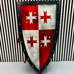 Może przedstawiać: Tarcza herbowa z ćwiartkowym wzorem w kolorach czerwonym i złamanej bieli. Każda sekcja zawiera stylizowany emblemat krzyża. Tarcza jest otoczona czarną ramą z równomiernie rozmieszczonymi srebrnymi nitami wzdłuż krawędzi.