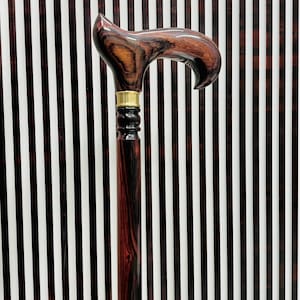 Puede incluir: Un bastón de madera con un mango curvo y un acabado marrón oscuro y rojo. El mango tiene un collar de latón y un diseño decorativo. El bastón está sobre un fondo a rayas.