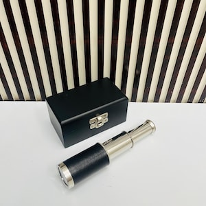 Puede incluir: Una caja de madera negra con un cierre plateado está junto a un telescopio plateado y negro. El telescopio tiene un agarre de cuero negro y está extendido. El fondo tiene un patrón de rayas marrones y blancas.