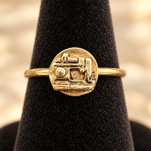 May include: A gold-toned ring featuring a circular face with a raised, detailed relief of a vintage sewing machine. The band is thin and smooth, and the entire piece has a matte, textured finish consistent with a cast metal construction.
