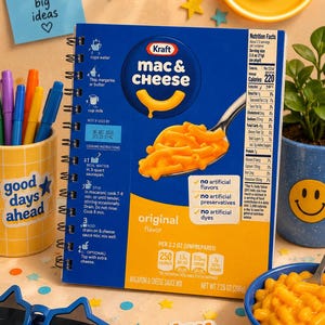 May include: A spiral-bound notebook featuring the blue and yellow Kraft Mac & Cheese box design on the cover. The cover displays cooking instructions, nutrition facts, and a graphic of a spoon holding macaroni. It measures 7.25 ounces.