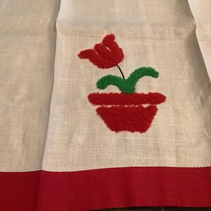 May include: A white linen-textured towel featuring a red border at the bottom and a central tufted embroidery design. The design depicts a red tulip flower with green leaves growing out of a red flower pot, rendered in a textured punch needle style.
