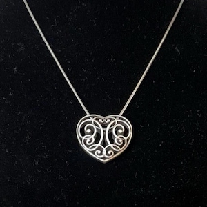 Puede incluir: Un collar con colgante de coraz&oacute;n plateado. El colgante en forma de coraz&oacute;n presenta un dise&ntilde;o calado abierto con patrones arremolinados. El collar tiene una delicada cadena plateada, sobre un fondo negro.