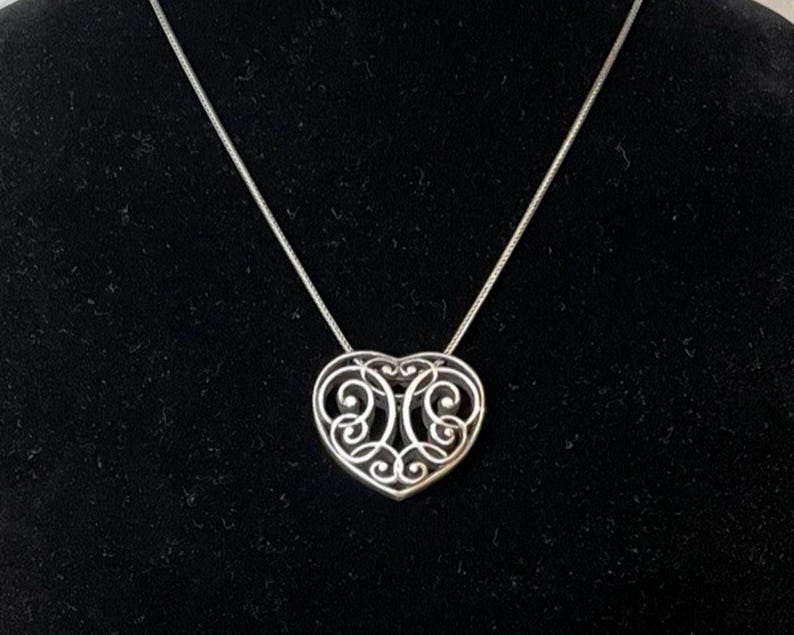 Puede incluir: Un collar con colgante de coraz&oacute;n plateado. El colgante en forma de coraz&oacute;n presenta un dise&ntilde;o calado abierto con patrones arremolinados. El collar tiene una delicada cadena plateada, sobre un fondo negro.