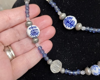 May include: A necklace featuring blue and white porcelain beads with a floral design, interspersed with silver-toned beads and small blue beads. The necklace is displayed against a dark background.