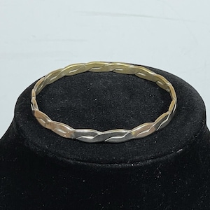 May include: A circular bangle bracelet featuring a twisted design. The metal construction alternates between sections of polished gold-tone and silver-tone finishes, creating a continuous braided appearance throughout the entire circumference of the band.
