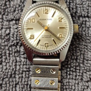 Peut inclure: Une montre-bracelet Mirexal vintage avec un boîtier argenté et un cadran doré. La montre présente des aiguilles et des index dorés, avec le texte "17 Jewels Incabloc" et "Swiss Made". La montre a un bracelet en métal argenté.
