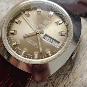 Puede incluir: Un reloj de pulsera Creation vintage con esfera dorada, bisel plateado y correa de cuero marrón. La esfera del reloj muestra la fecha DIM 22 y el texto "AUTOMATIC INCABLOC". El reloj tiene un diseño clásico y retro.