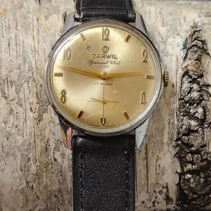 Może przedstawiać: Zegarek Darwil Special Flat Lux w stylu vintage ze złotą tarczą i czarnym skórzanym paskiem. Tarcza zegarka ma złote wskazówki i logo Darwil, z napisami "Special Flat Lux" i "17 Rubis". Zegarek jest prezentowany na teksturowanym tle.