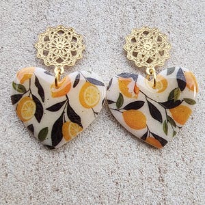 Peut inclure: Boucles d'oreilles en forme de cœur avec un motif de citron. Elles sont composées d'une partie supérieure en filigrane dorée et d'un pendentif en forme de cœur avec un fond crème, des citrons oranges et des feuilles vert foncé. Un accessoire unique.