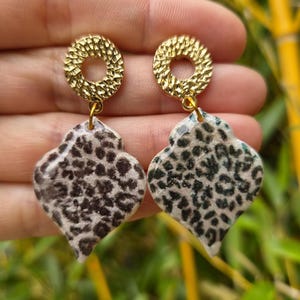 May include: A pair of drop earrings featuring a gold-toned circular stud with a textured, hammered finish. Attached to each stud is a dangling, teardrop-shaped pendant with a smooth, glossy surface displaying a black and beige leopard print pattern.