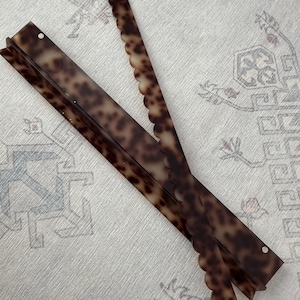 May include: Two long, narrow hair accessories with a brown and black tortoiseshell pattern. One piece features a straight edge, while the other piece has a decorative scalloped edge. Both items are thin, flat, and rectangular in shape.