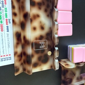 May include: A tortoise shell patterned acrylic rack for game tiles with a scalloped edge and a white logo featuring the letters M and M. The rack holds several solid pink rectangular tiles and a stack of multi-colored tiles in pink, yellow, and purple.
