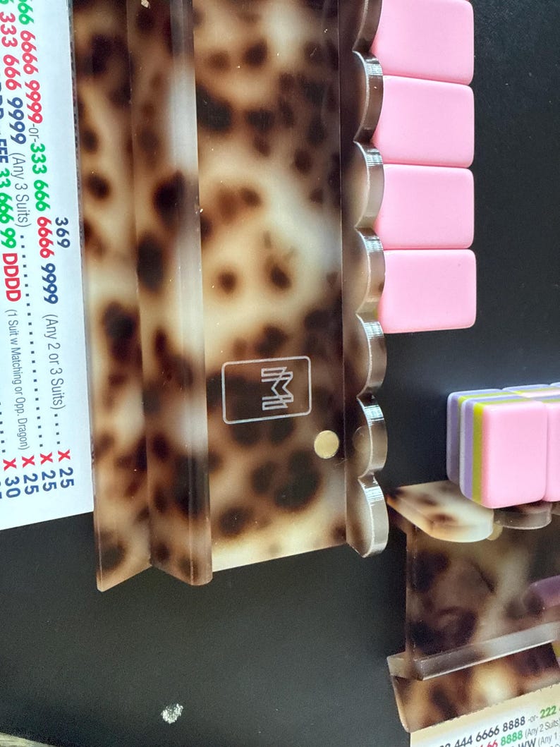 May include: A tortoise shell patterned acrylic rack for game tiles with a scalloped edge and a white logo featuring the letters M and M. The rack holds several solid pink rectangular tiles and a stack of multi-colored tiles in pink, yellow, and purple.
