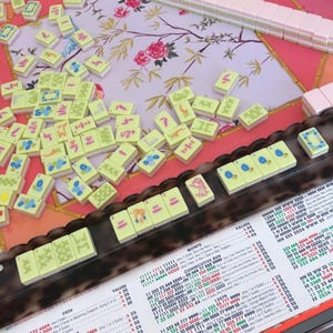 May include: A Mah Jongg set featuring light green tiles with various blue, red, and black symbols, arranged on a pink floral mat. The set includes a dark brown rack, a white score card with black text, and a stack of pink tiles on the side.