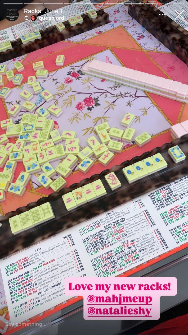 May include: A Mah Jongg set featuring light green tiles with various blue, red, and black symbols, arranged on a pink floral mat. The set includes a dark brown rack, a white score card with black text, and a stack of pink tiles on the side.