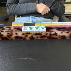May include: A tortoiseshell patterned Mahjong tile rack with a scalloped front edge, holding a row of pastel-colored Mahjong tiles. The tiles feature pink, yellow, and light blue horizontal stripes, with some displaying green bamboo designs and a purple flower.