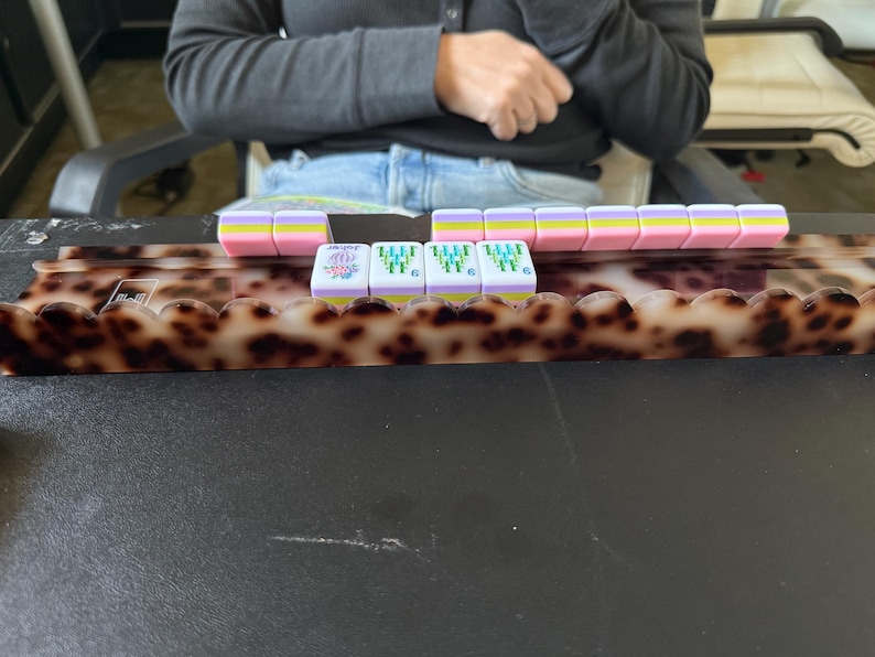 May include: A tortoiseshell patterned Mahjong tile rack with a scalloped front edge, holding a row of pastel-colored Mahjong tiles. The tiles feature pink, yellow, and light blue horizontal stripes, with some displaying green bamboo designs and a purple flower.