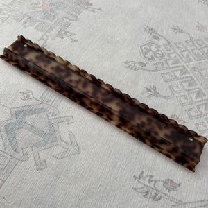 May include: A long, rectangular hair accessory featuring a brown and black tortoiseshell pattern. The item has a scalloped top edge and a small circular hole at each end, with a smooth, glossy finish throughout its elongated, narrow structure.