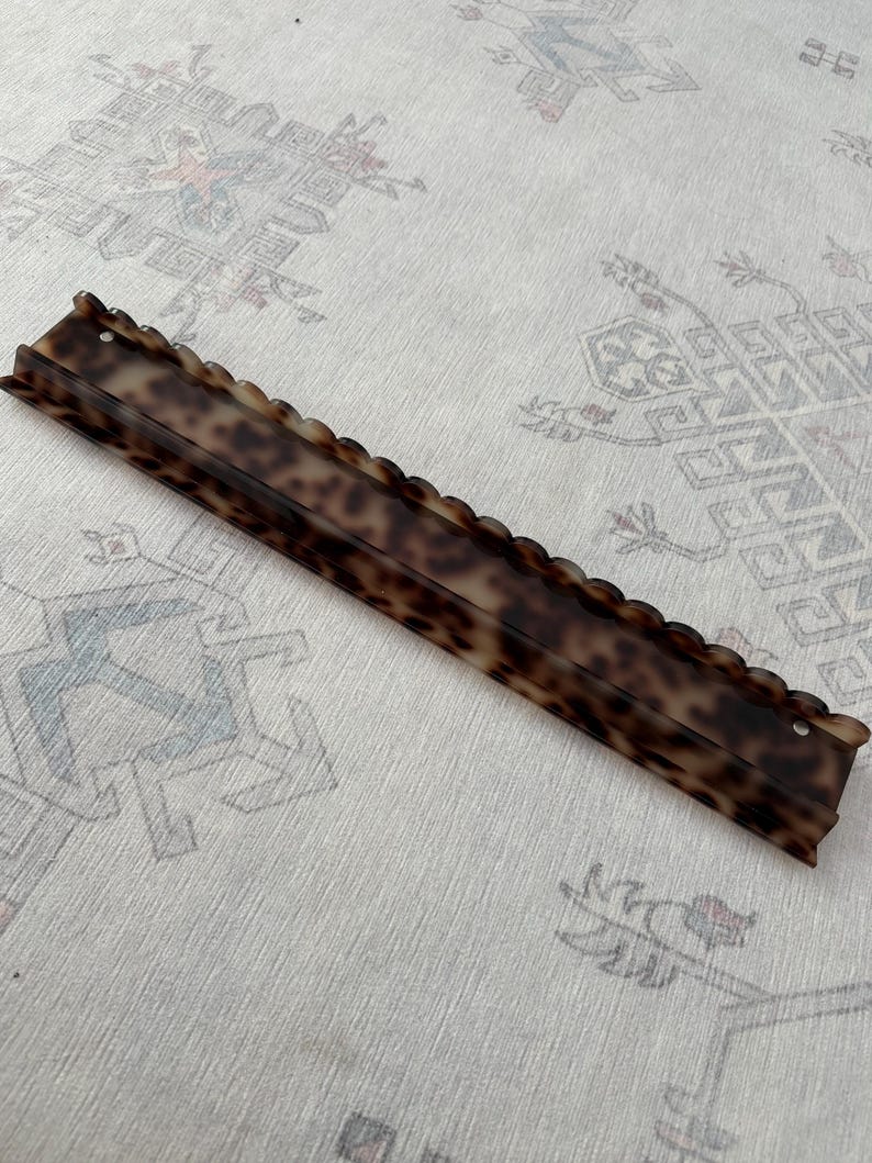 May include: A long, rectangular hair accessory featuring a brown and black tortoiseshell pattern. The item has a scalloped top edge and a small circular hole at each end, with a smooth, glossy finish throughout its elongated, narrow structure.