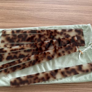 Peut inclure: Un ensemble de quatre bâtonnets à cheveux rectangulaires avec un motif écaille de tortue marron et crème, disposés en éventail sur une pochette à cordon en tissu vert sauge clair avec des liens blancs et des œillets dorés.