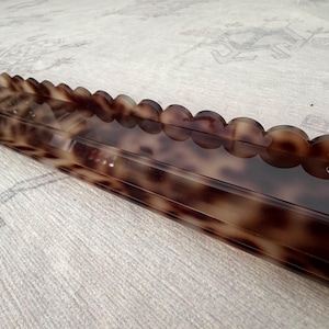 May include: A long, rectangular shelf featuring a glossy, brown and black tortoiseshell pattern. The front edge of the shelf is designed with a decorative scalloped trim, and a small circular hole is visible on the right side of the front panel.