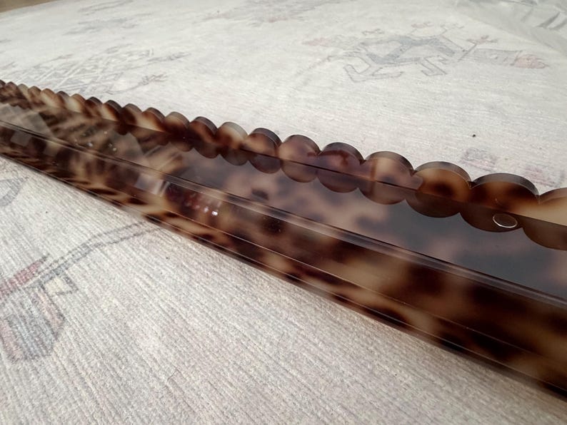 May include: A long, rectangular shelf featuring a glossy, brown and black tortoiseshell pattern. The front edge of the shelf is designed with a decorative scalloped trim, and a small circular hole is visible on the right side of the front panel.