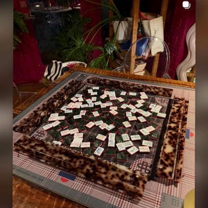 May include: A mahjong set featuring a square board with a brown and black tortoiseshell pattern frame. The game includes numerous white tiles with green backs and various red, blue, and black character designs, arranged on a plaid game mat.