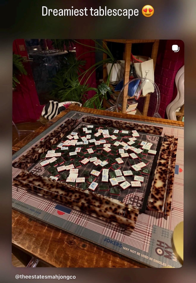 May include: A mahjong set featuring a square board with a brown and black tortoiseshell pattern frame. The game includes numerous white tiles with green backs and various red, blue, and black character designs, arranged on a plaid game mat.