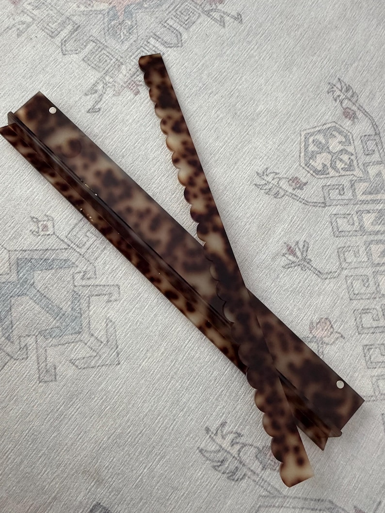 May include: Two long, narrow hair accessories with a brown and black tortoiseshell pattern. One piece features a straight edge, while the other piece has a decorative scalloped edge. Both items are thin, flat, and rectangular in shape.