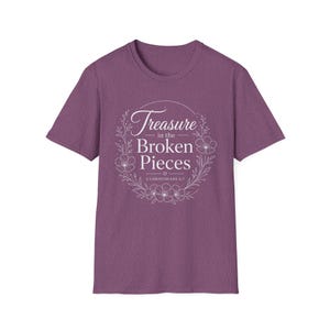May include: A heathered purple short-sleeve t-shirt featuring a white graphic design of a floral wreath surrounding the text "Treasure in the Broken Pieces" and "2 Corinthians 4:7" printed in a mix of script and serif fonts on the chest.