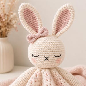 May include: A cream-colored crochet bunny lovey featuring long ears with pink inner sections, a small pink bow attached to the side of the head, and embroidered closed eyes with a black nose. The face has soft pink blush accents on the cheeks.
