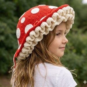 May include: A crochet bucket hat featuring a bright red dome with white circular spots, resembling a mushroom cap. The hat has a wide, ruffled cream-colored brim with a textured, wavy edge that frames the face, crafted from yarn.